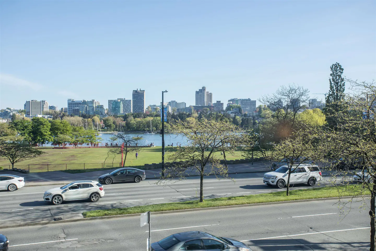 View of a city skyline over a lake and park, with cars on a road in the foreground on a sunny day.