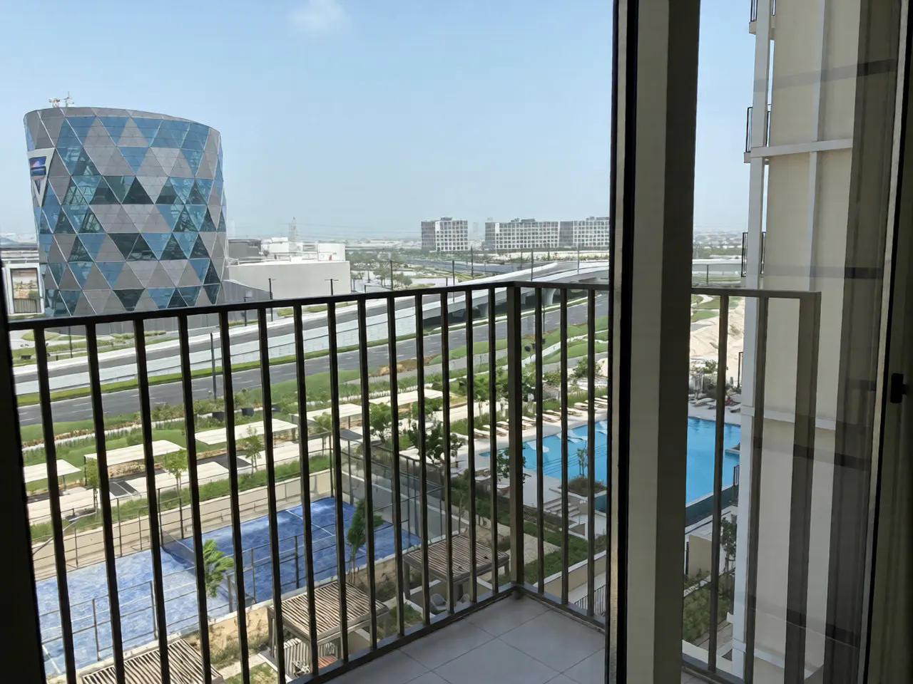 View from a balcony with black railings overlooking a pool, tennis court, and a modern building in the distance.