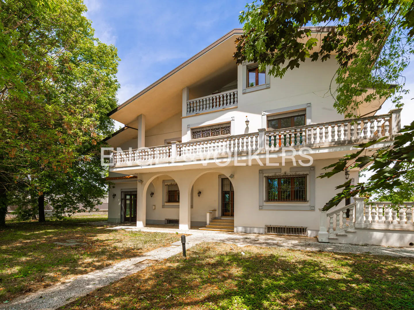 A three-story cream-colored villa with arched ground-floor entryways, stone balustrades, and a large, tree-lined yard.