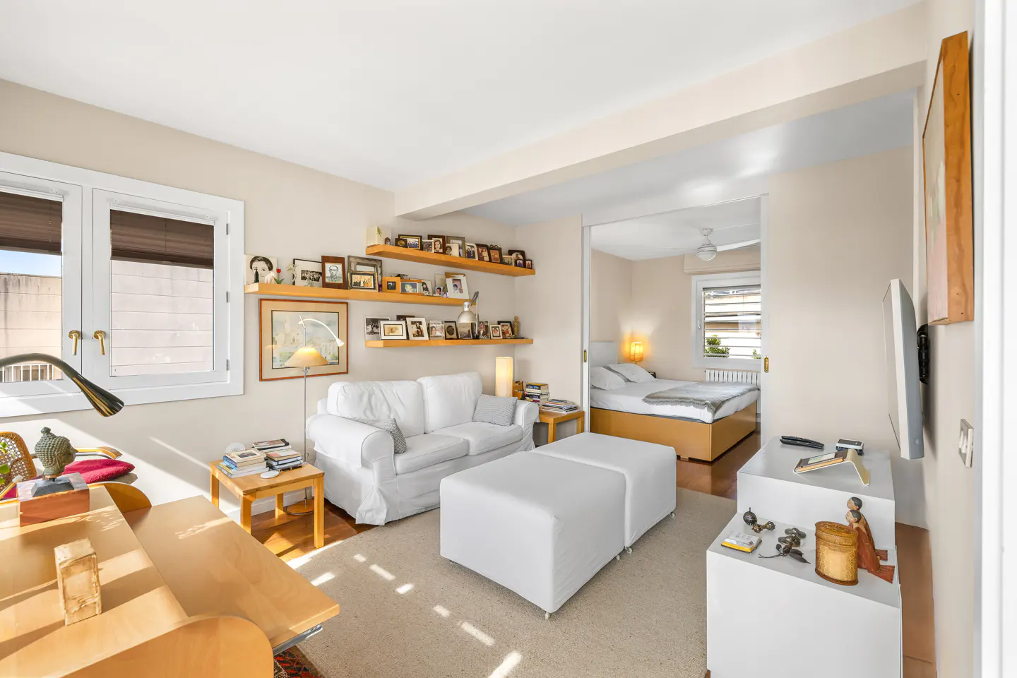 Bright apartment living room with white sofa, ottoman, and shelves of photos. Bedroom visible through doorway.