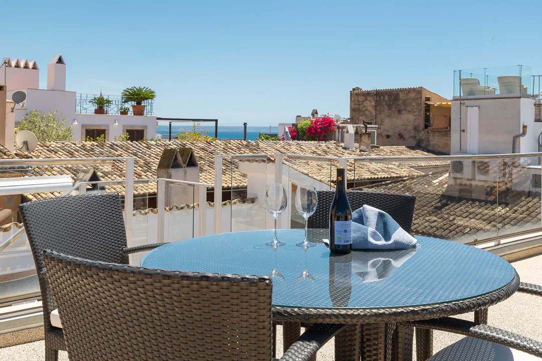 Rooftop patio with a blue glass table, wine bottle, glasses, and wicker chairs overlooking terracotta rooftops and the sea.