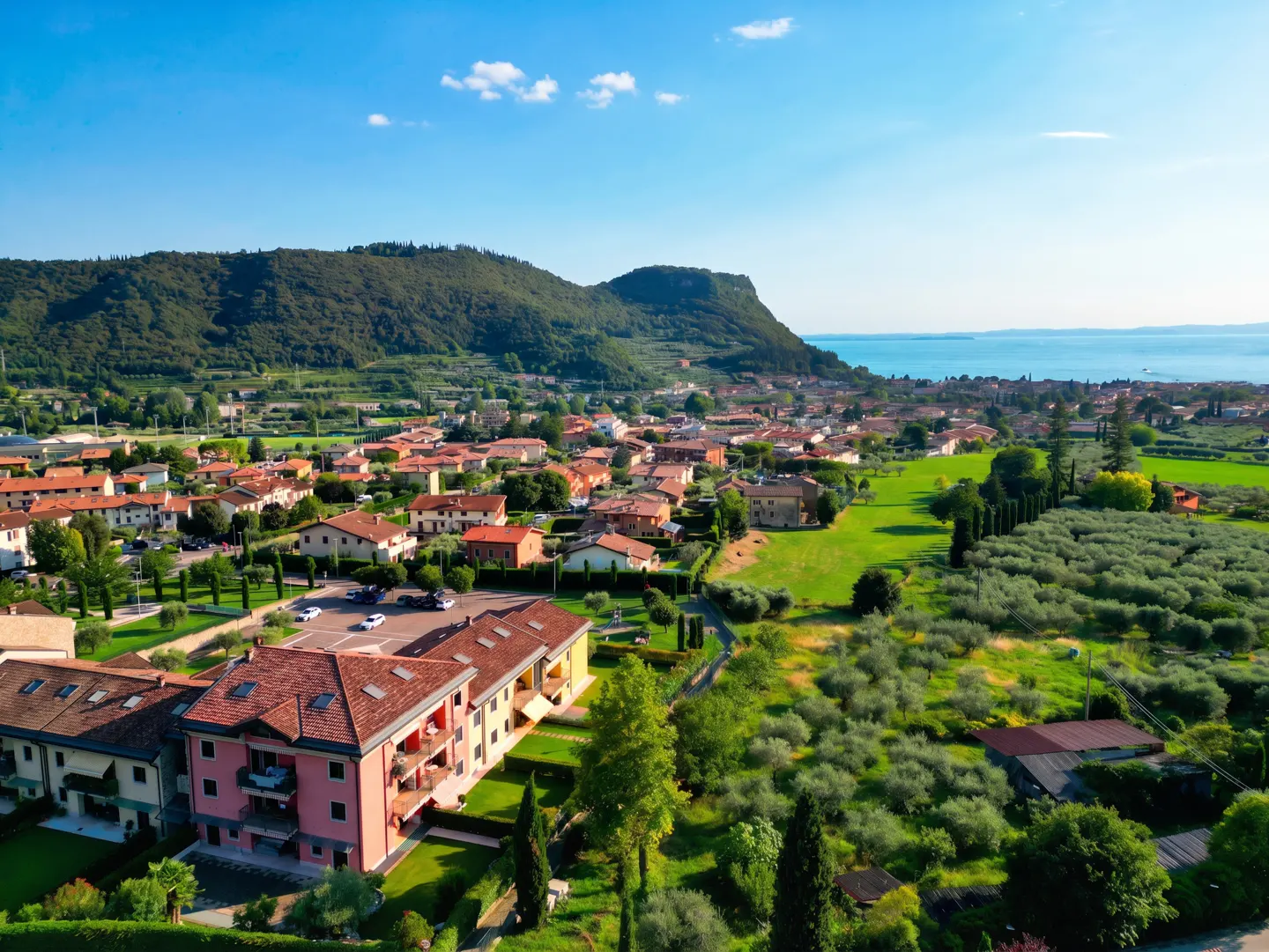 Aerial view of a town with red-roofed buildings, green fields, and a mountain backdrop under a blue sky.
