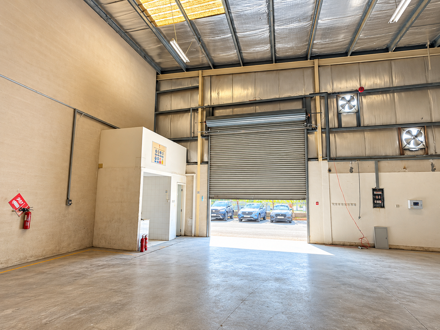 Empty industrial warehouse with concrete floors, high ceilings, a roll-up door, and a small office area with fire extinguishers. [AI-generated text]