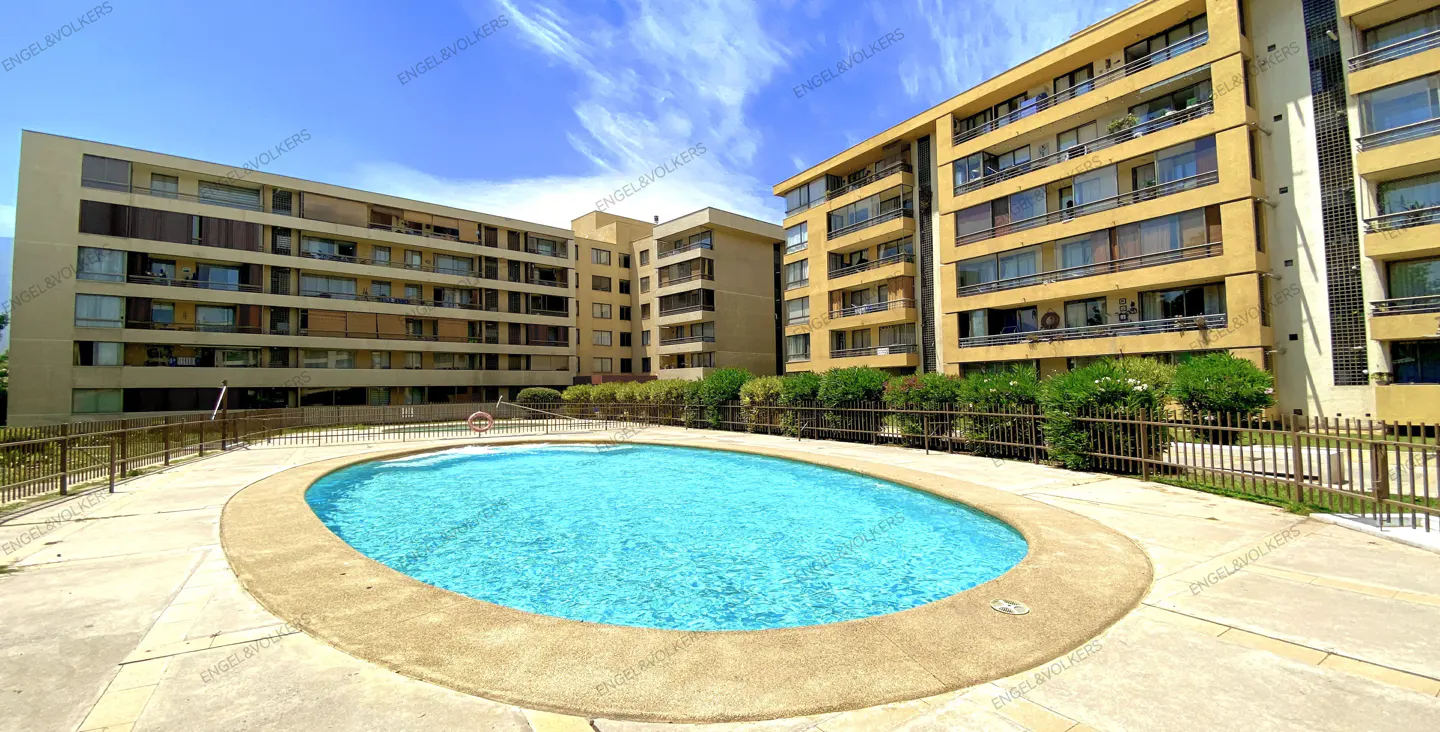 Outdoor pool with turquoise water in front of tan apartment buildings under a blue sky.