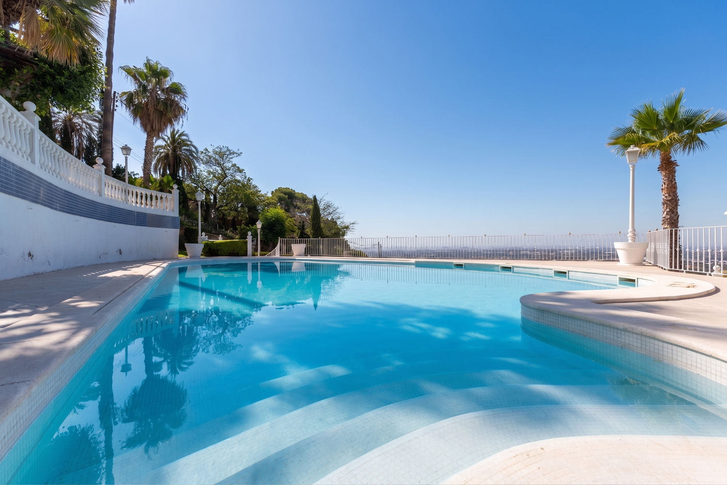 A bright blue swimming pool with steps, surrounded by palm trees and a white railing against a clear sky. [AI-generated text]
