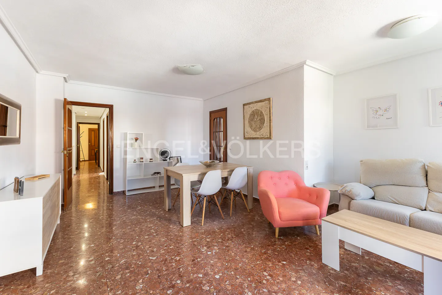Bright living room with white walls, red speckled floor, dining table, pink chair, and beige sofa.