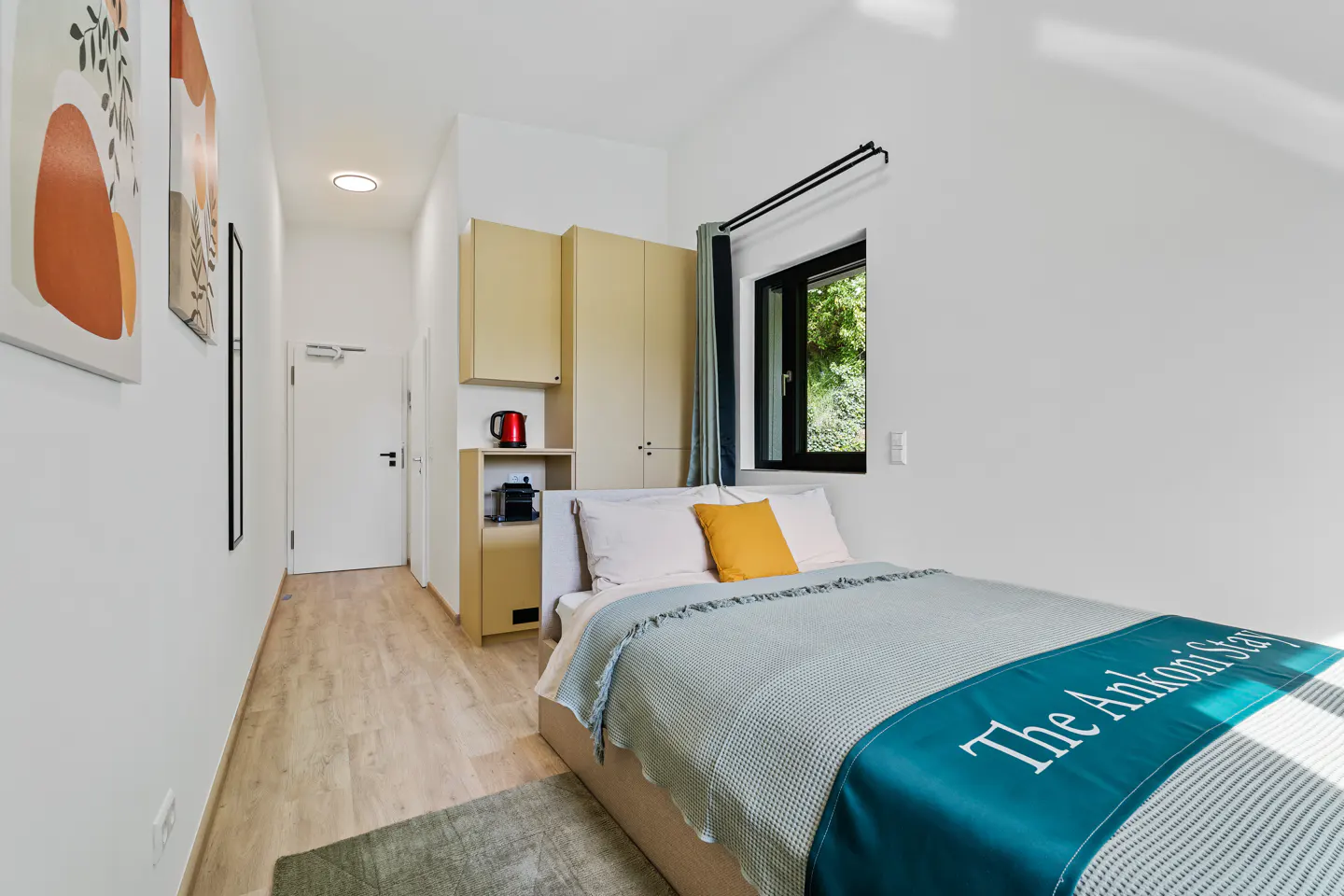 Bright bedroom with white walls, light wood floors, and a bed with a blue blanket. A window and storage unit are visible.