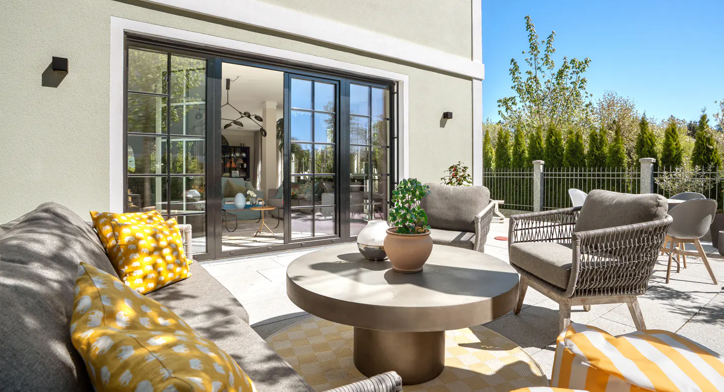 Outdoor patio with gray sofa, yellow pillows, round table, and chairs. Black framed sliding glass doors lead to the interior living space.