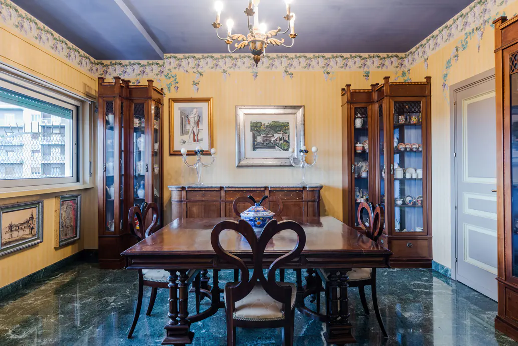 A dining room with a dark wood table, chairs, cabinets, and marble floor. The walls are yellow with a floral border, and a chandelier hangs from the blue ceiling.