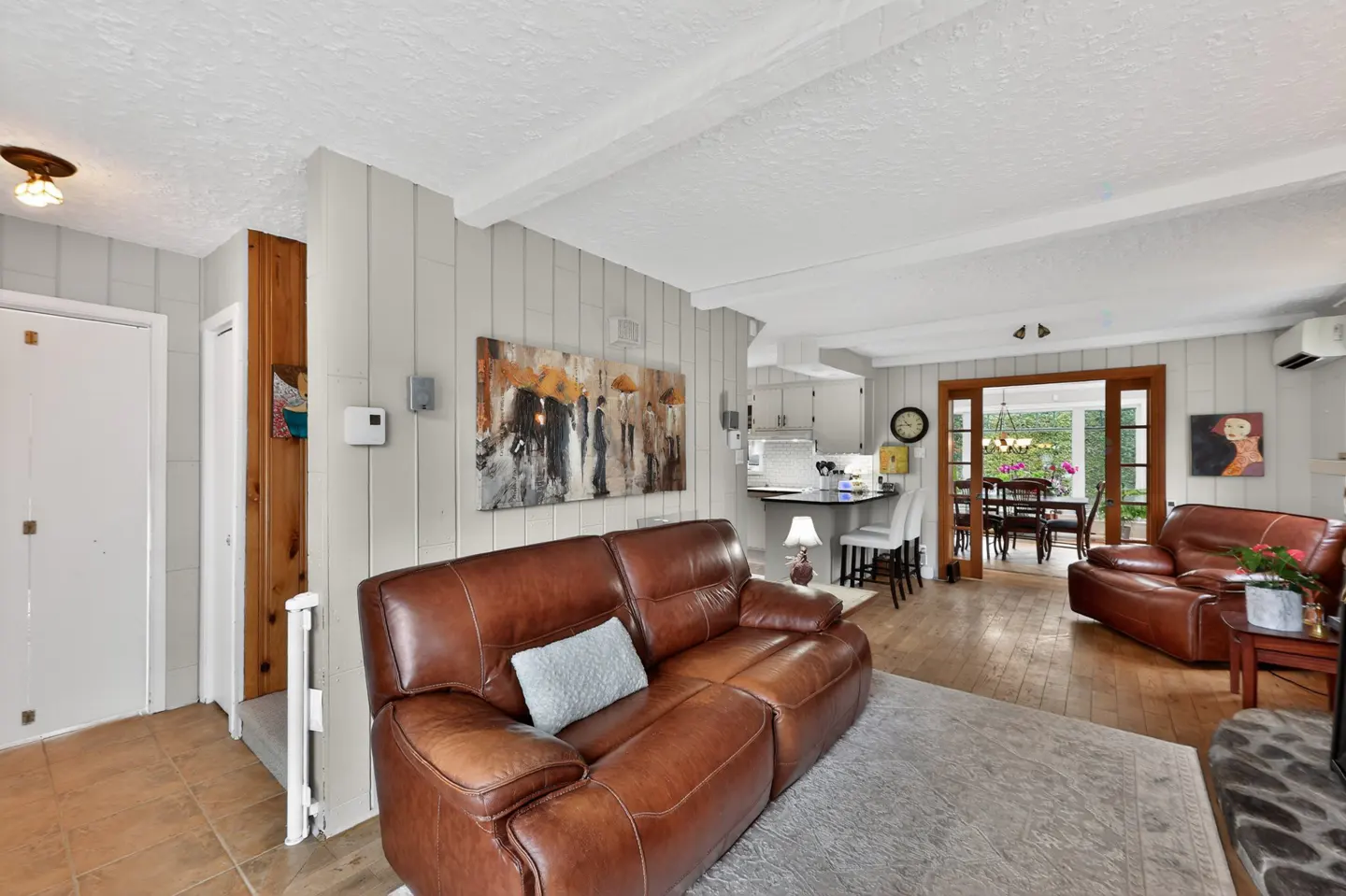 Living room with brown leather sofas, gray walls, and wood floors. A painting hangs above the sofa. A dining room is visible through a doorway.