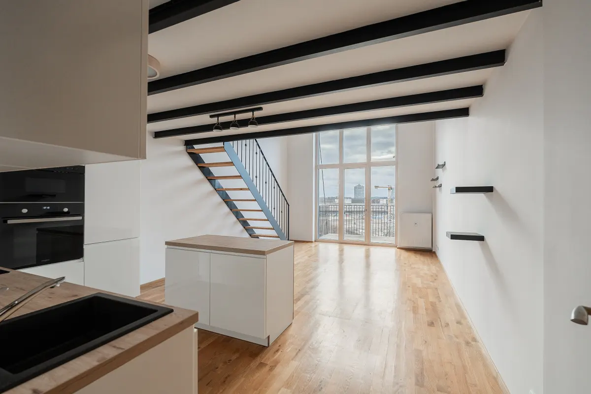 Bright, modern apartment interior with white walls, wood floors, black ceiling beams, and a staircase leading to a large window.