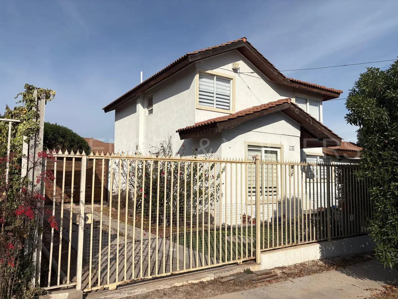 Two-story white house with a brown tile roof behind a tall, beige metal fence on a sunny day.