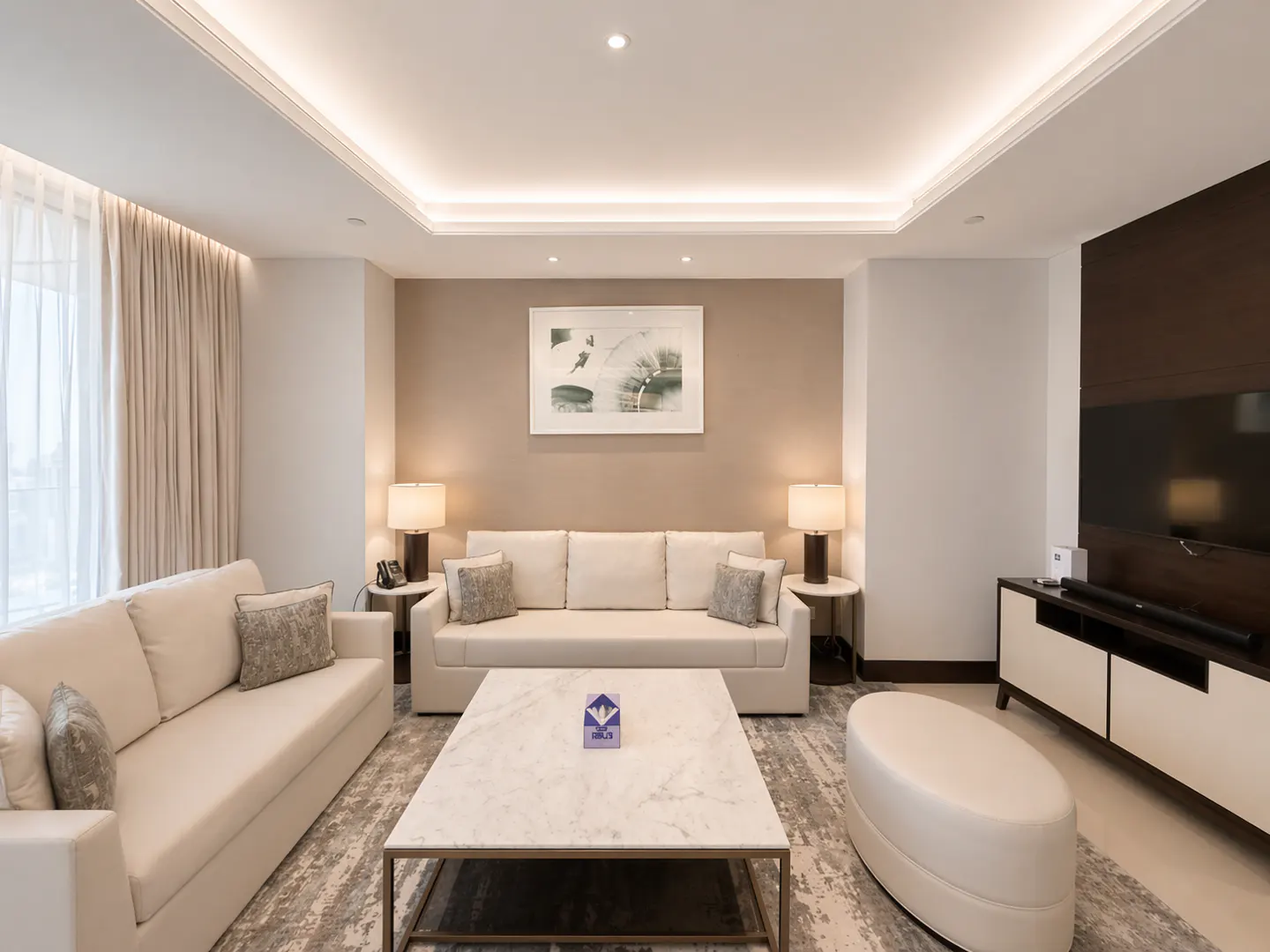 Living room with two white sofas, marble coffee table, and TV. Neutral tones, lamps, and artwork create a cozy atmosphere.