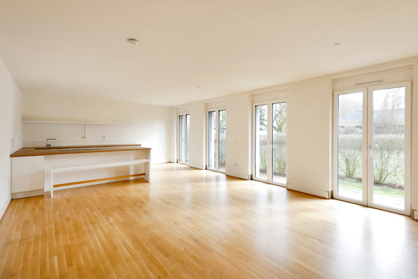 Bright, empty room with wood floors, white walls, and a kitchen counter. Three sets of glass doors lead to the outside.