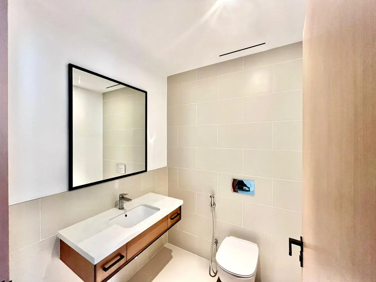 Modern bathroom with white tiles, toilet, and sink with a black-framed mirror above a wood vanity.