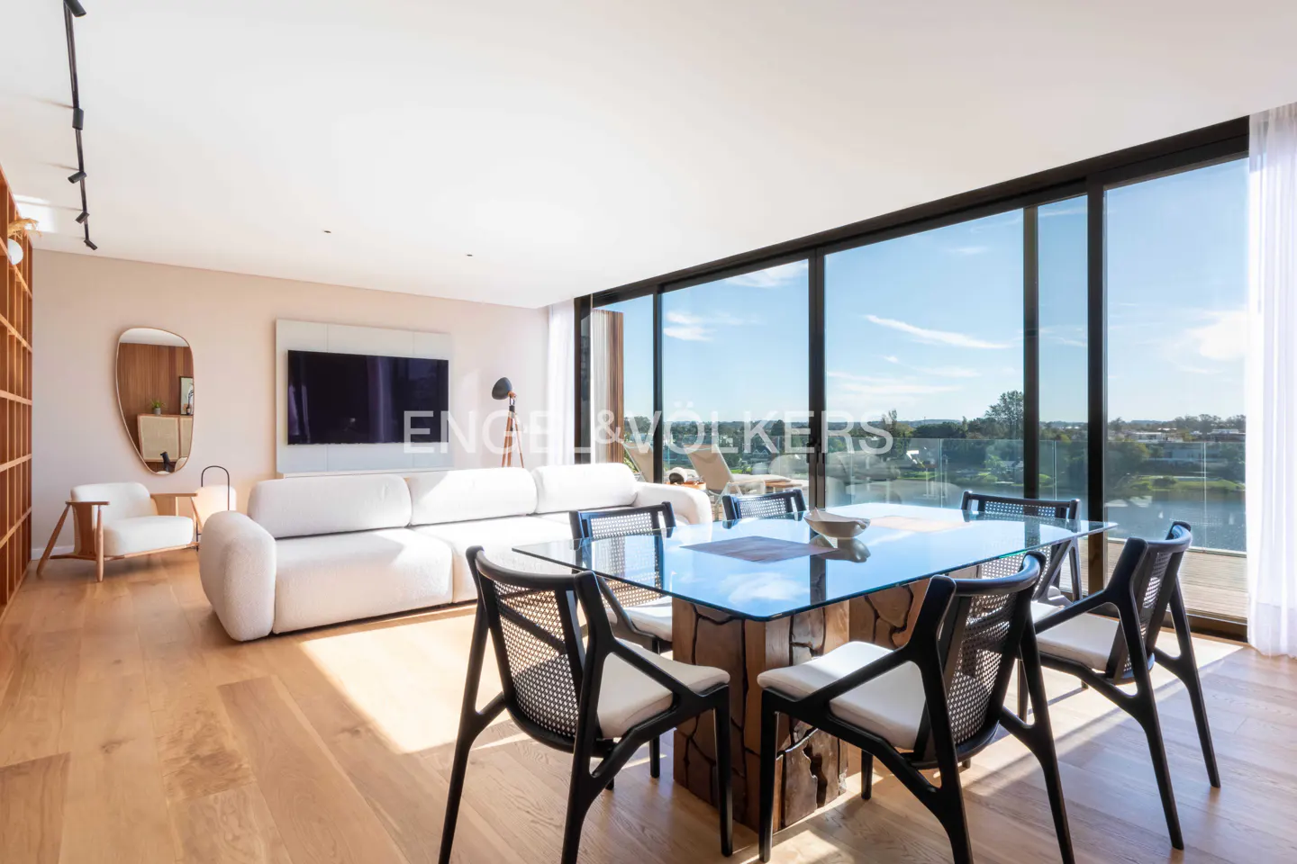 Modern living room with white sofa, glass dining table, black chairs, and floor-to-ceiling windows overlooking a landscape.