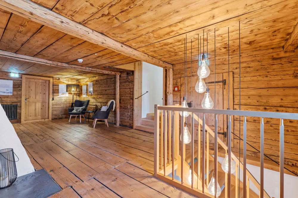 Rustic interior with wood floors, walls, and ceiling. A staircase with metal railing and hanging lightbulbs is on the right. Two chairs are on the left.