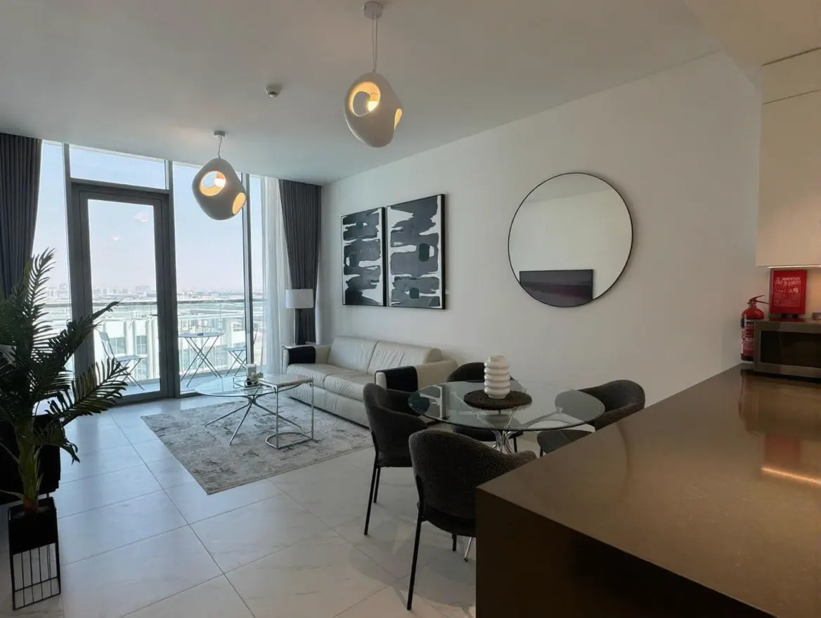 Bright, modern apartment with a white sofa, glass table, and city view from the balcony. Black and white art adorns the walls.