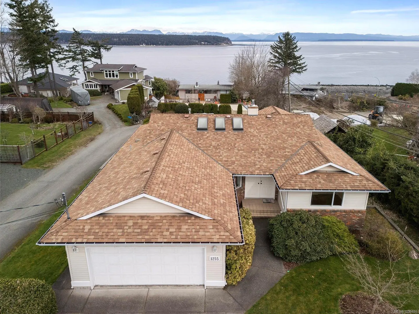 Aerial view of a one-story house with a brown roof, white garage door, and ocean view.