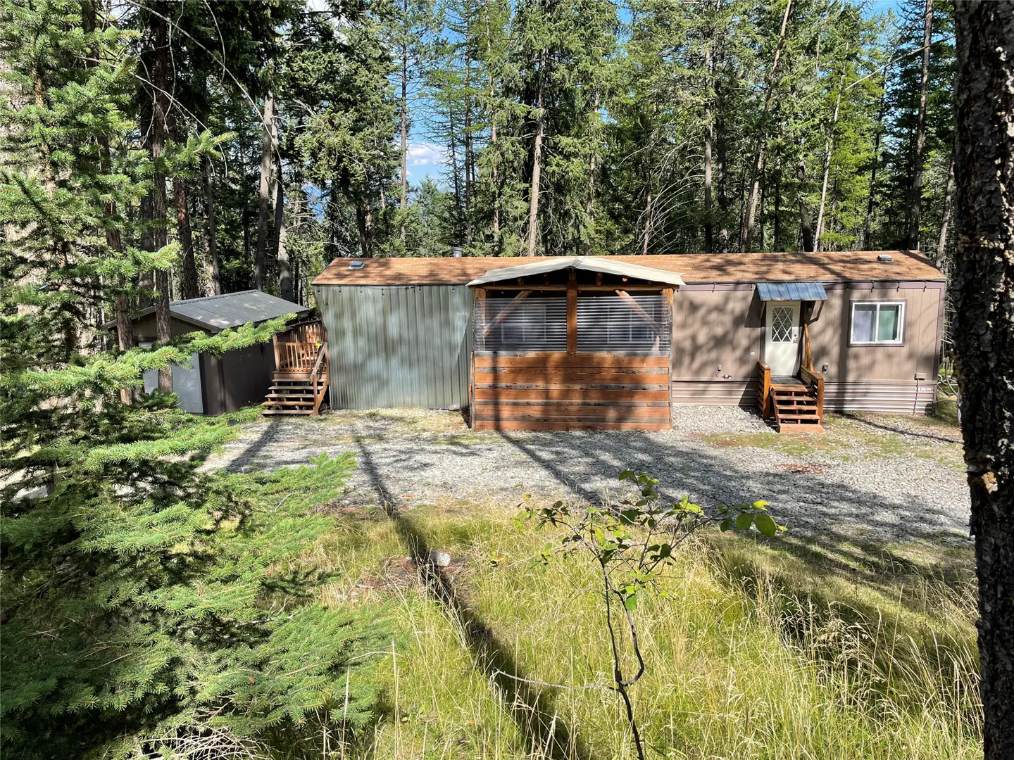 A brown mobile home with a porch and shed sits in a wooded area with tall trees.