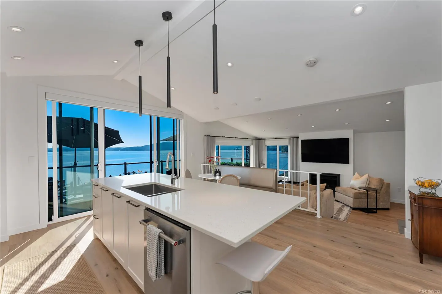 Open-concept kitchen and living room with a view of the ocean. White cabinets, island, and walls. Wood floors and black pendant lights.