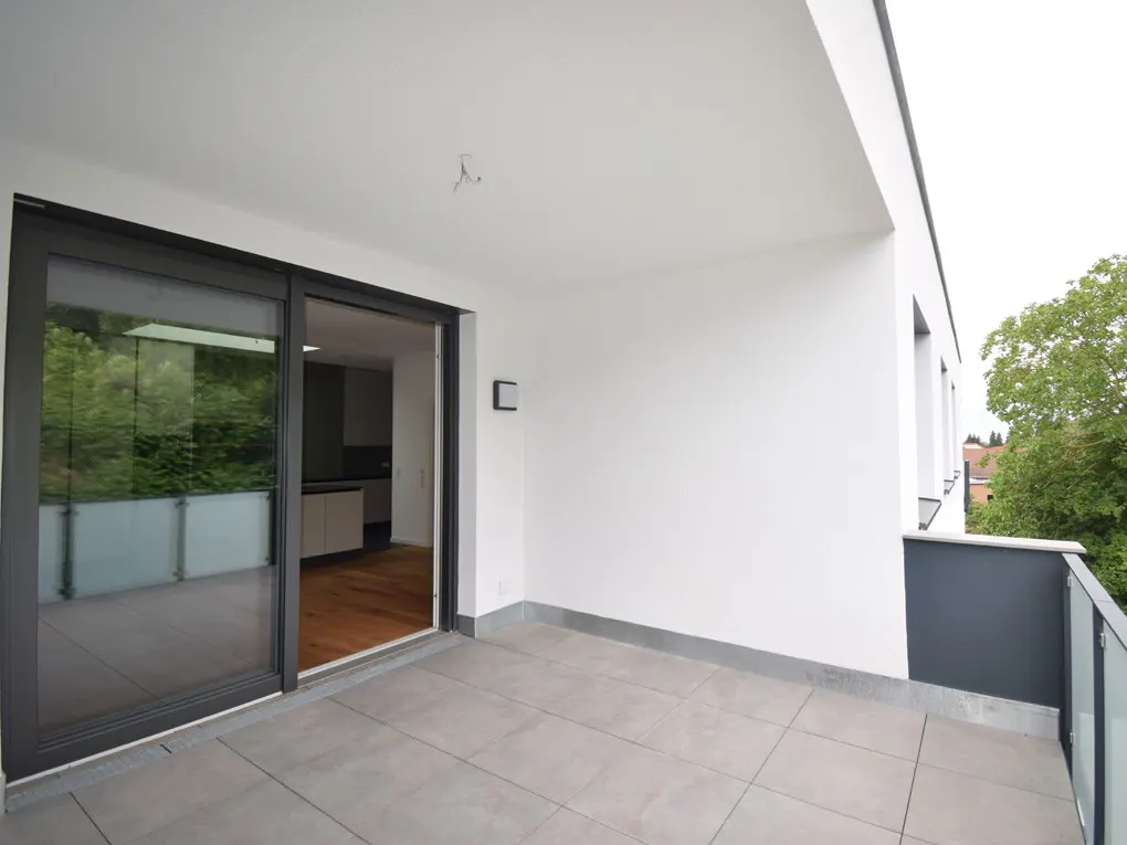 A modern balcony with gray tiles, a glass railing, and a sliding glass door leading to a kitchen with wood floors.