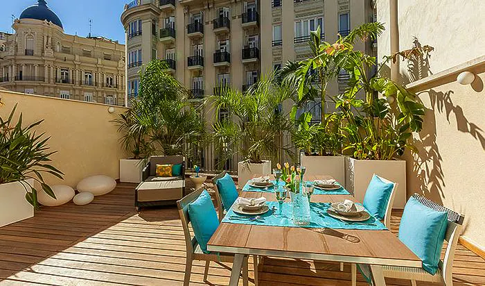 Outdoor dining area on a wooden deck with a table set for six, turquoise accents, potted plants, and city views.