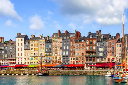 Waterfront view of Honfleur, France, featuring colorful buildings, boats, and awnings under a blue sky with clouds.