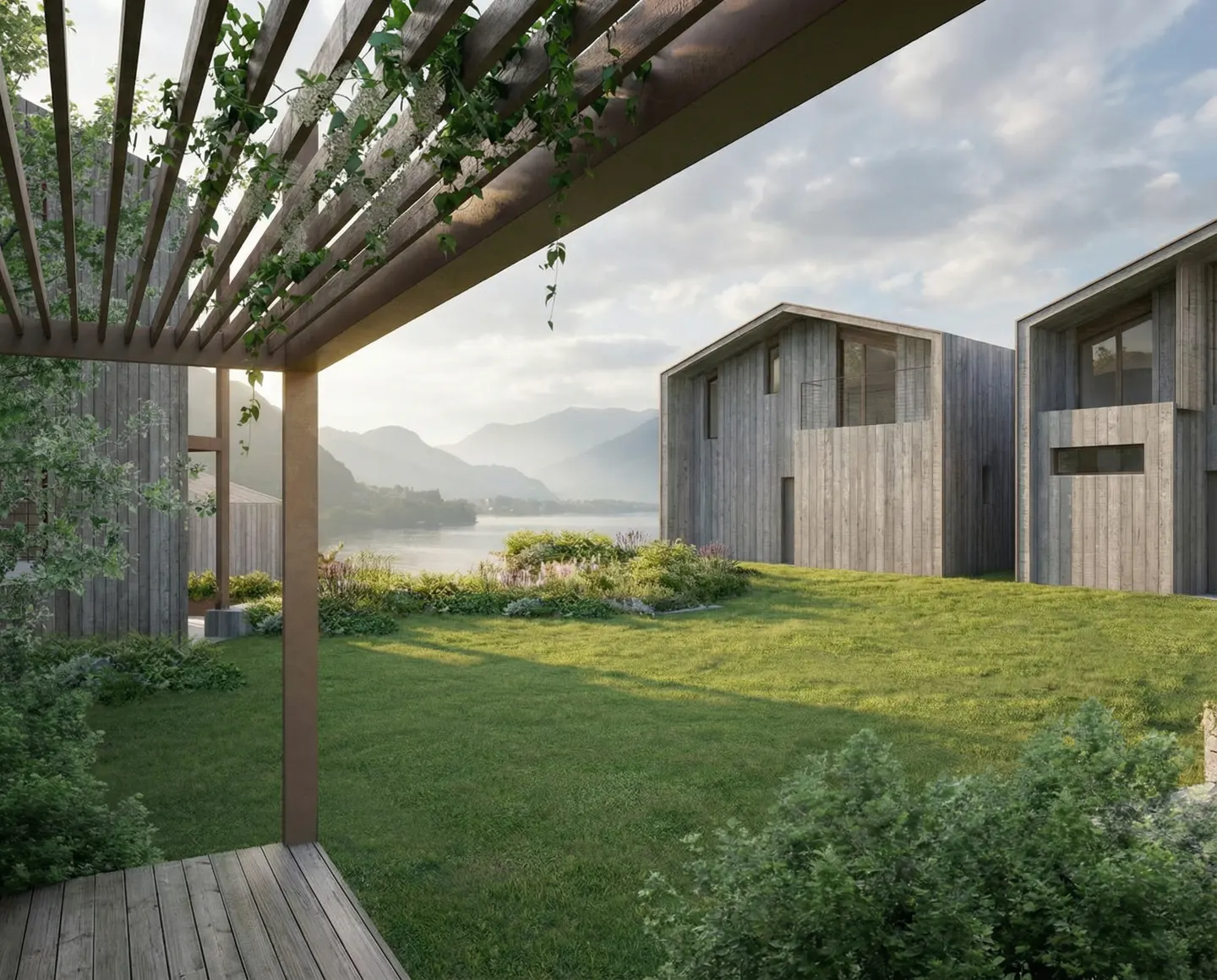 View from a wooden pergola onto a green lawn, modern wood houses, and a lake with mountains in the background.