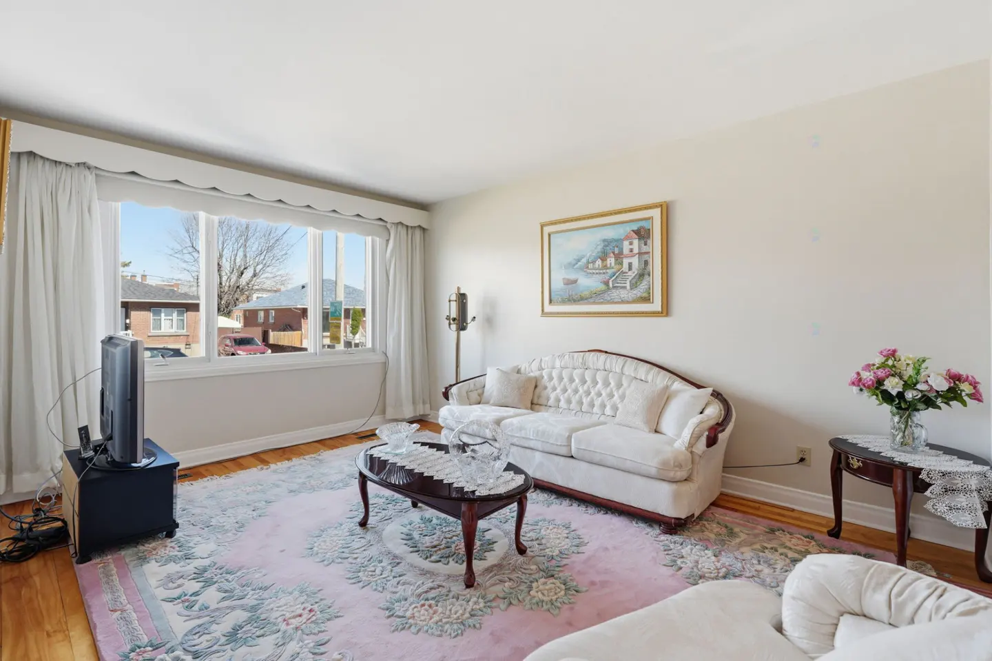 Living room with white sofa, floral rug, dark wood tables, and a large window with white curtains.