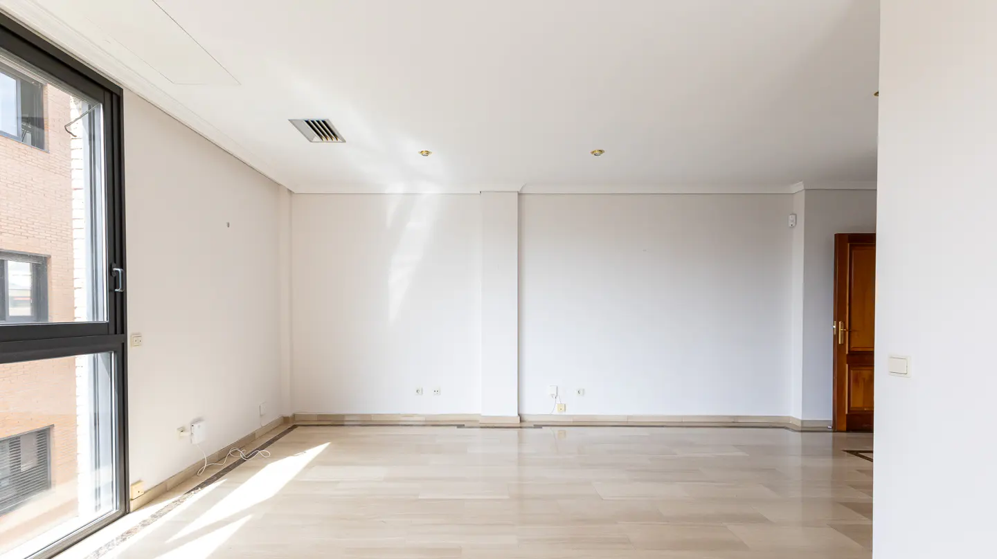 Bright, empty room with white walls, light wood floors, and a large black-framed window showing a brick building outside. A wooden door is visible on the right.