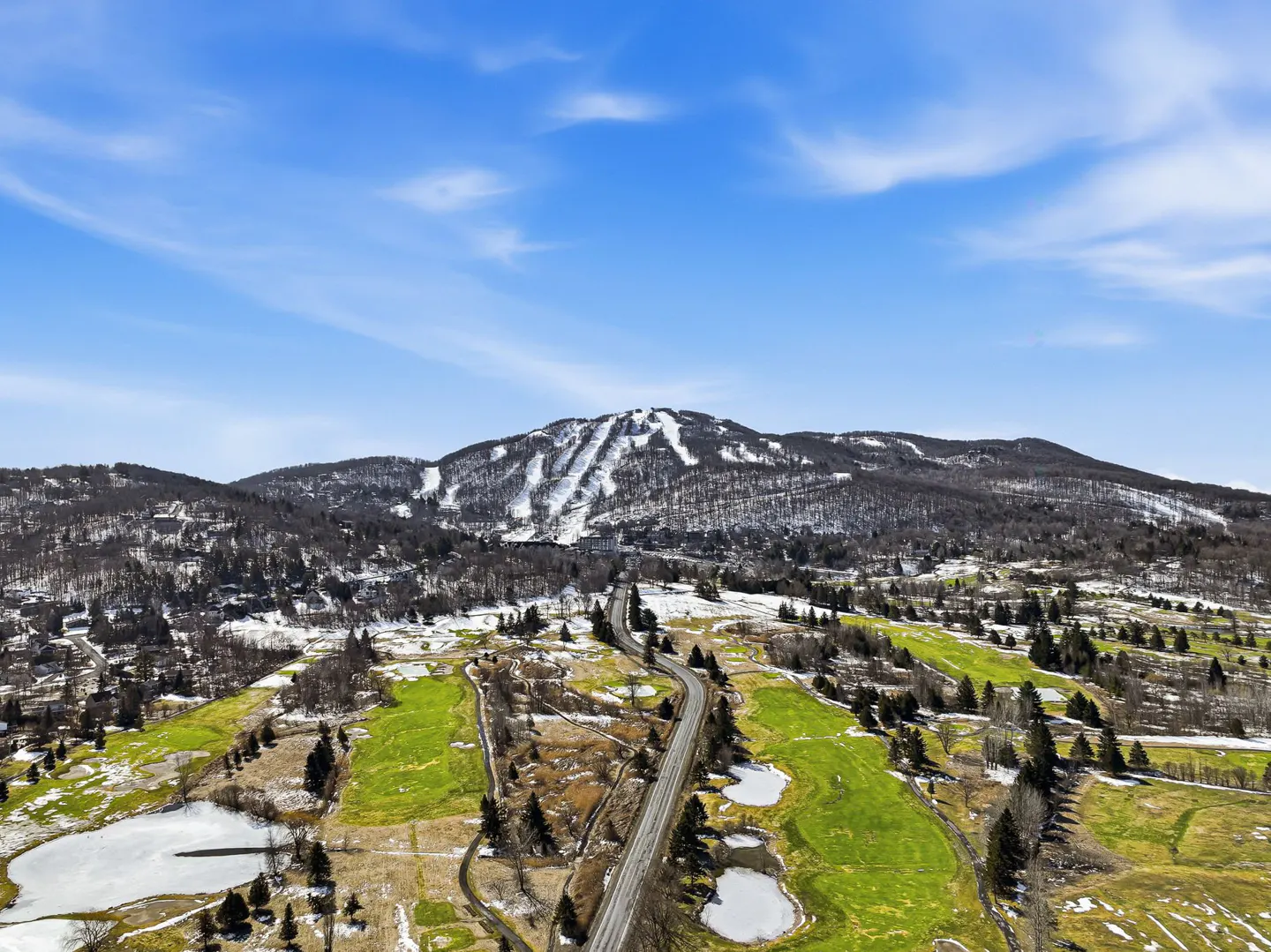 Scenic view of a mountain with ski slopes, green fields, and a winding road under a blue sky.