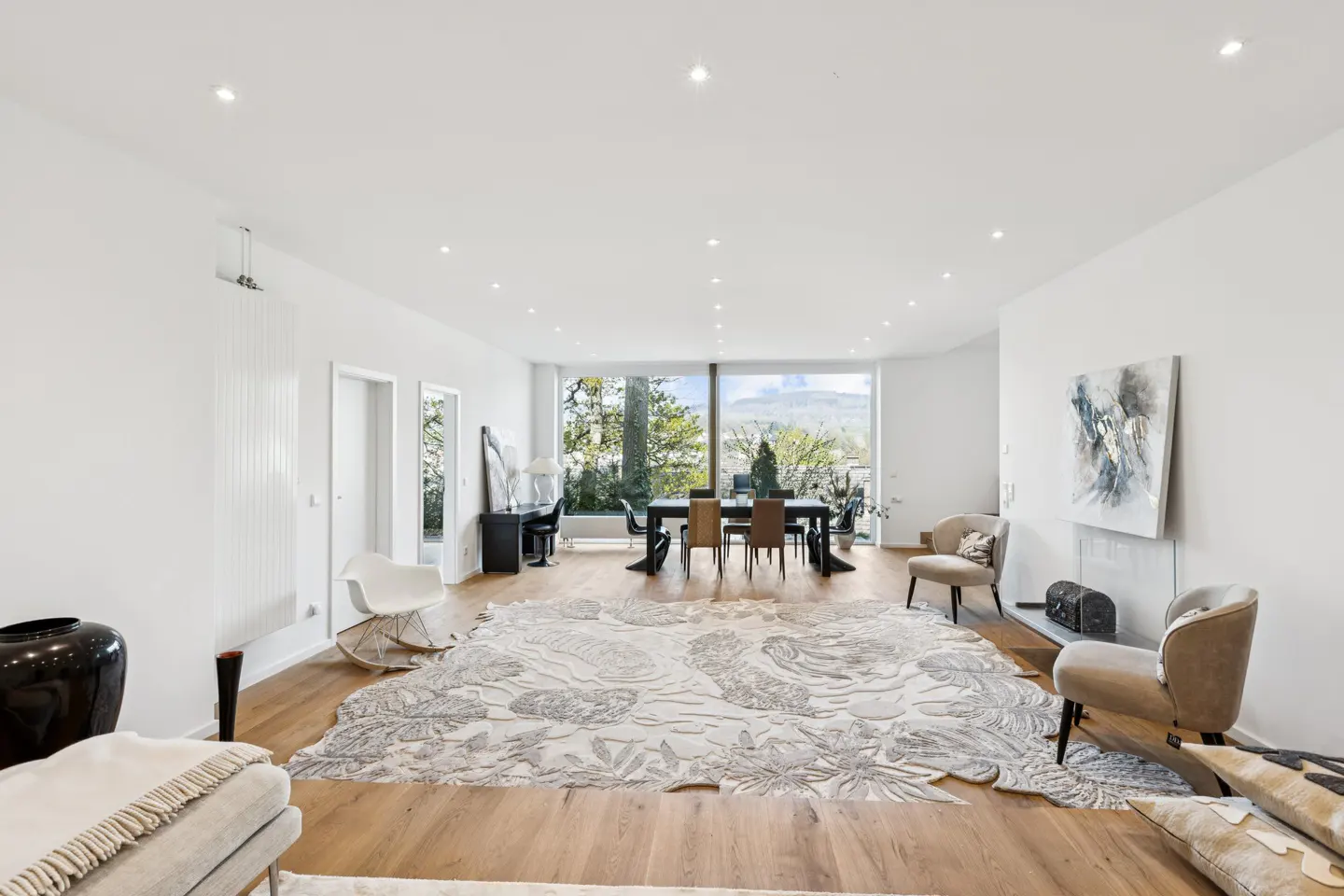 Bright, open-plan living and dining area with white walls, wood floors, and a large floral rug. A dining table sits in front of a large window with a view of trees and mountains.