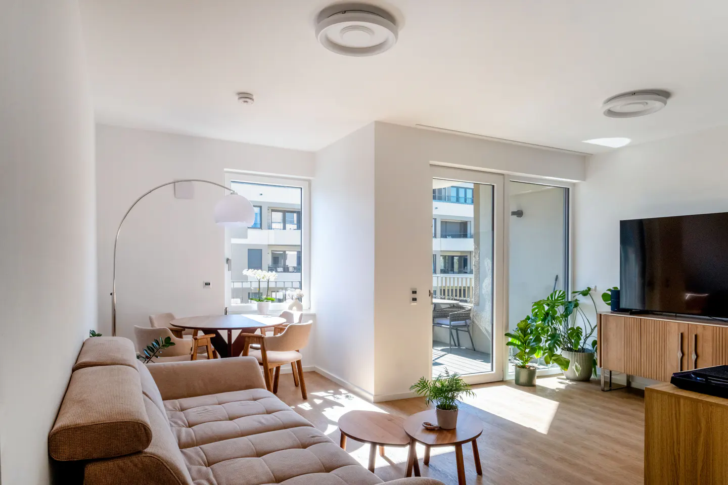 Bright living room with a beige sofa, wooden coffee tables, dining area, TV console, and balcony access with plants.