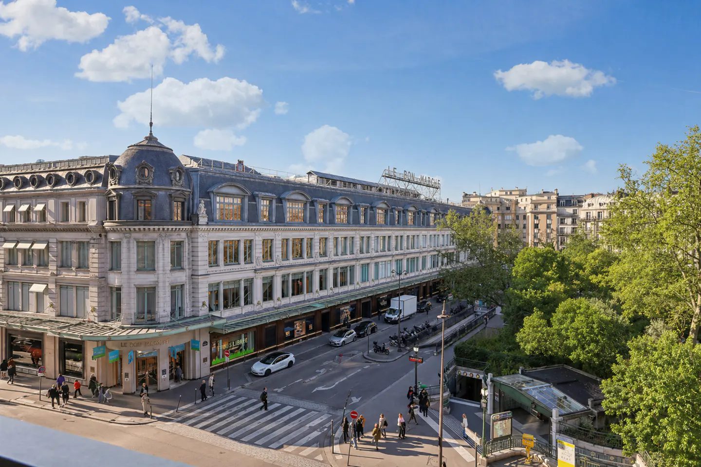 Paris street view of Le Bon Marché department store with crosswalk, cars, and people on a sunny day.