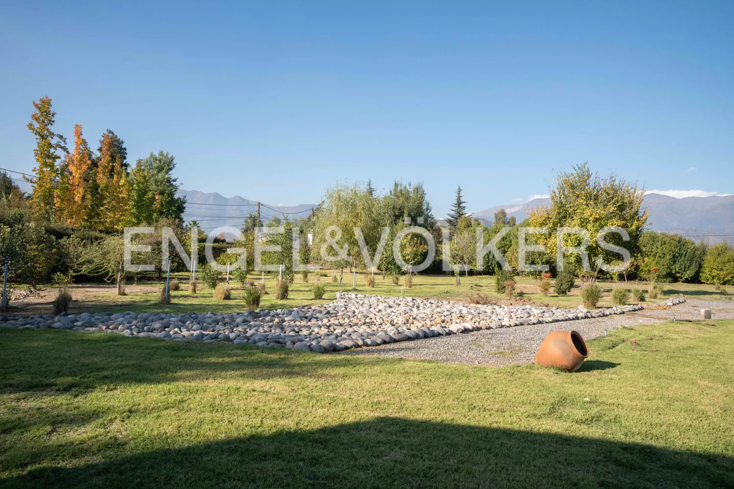 Lush green lawn with trees, rocks, and a clay pot. Mountains are visible in the background under a clear blue sky.