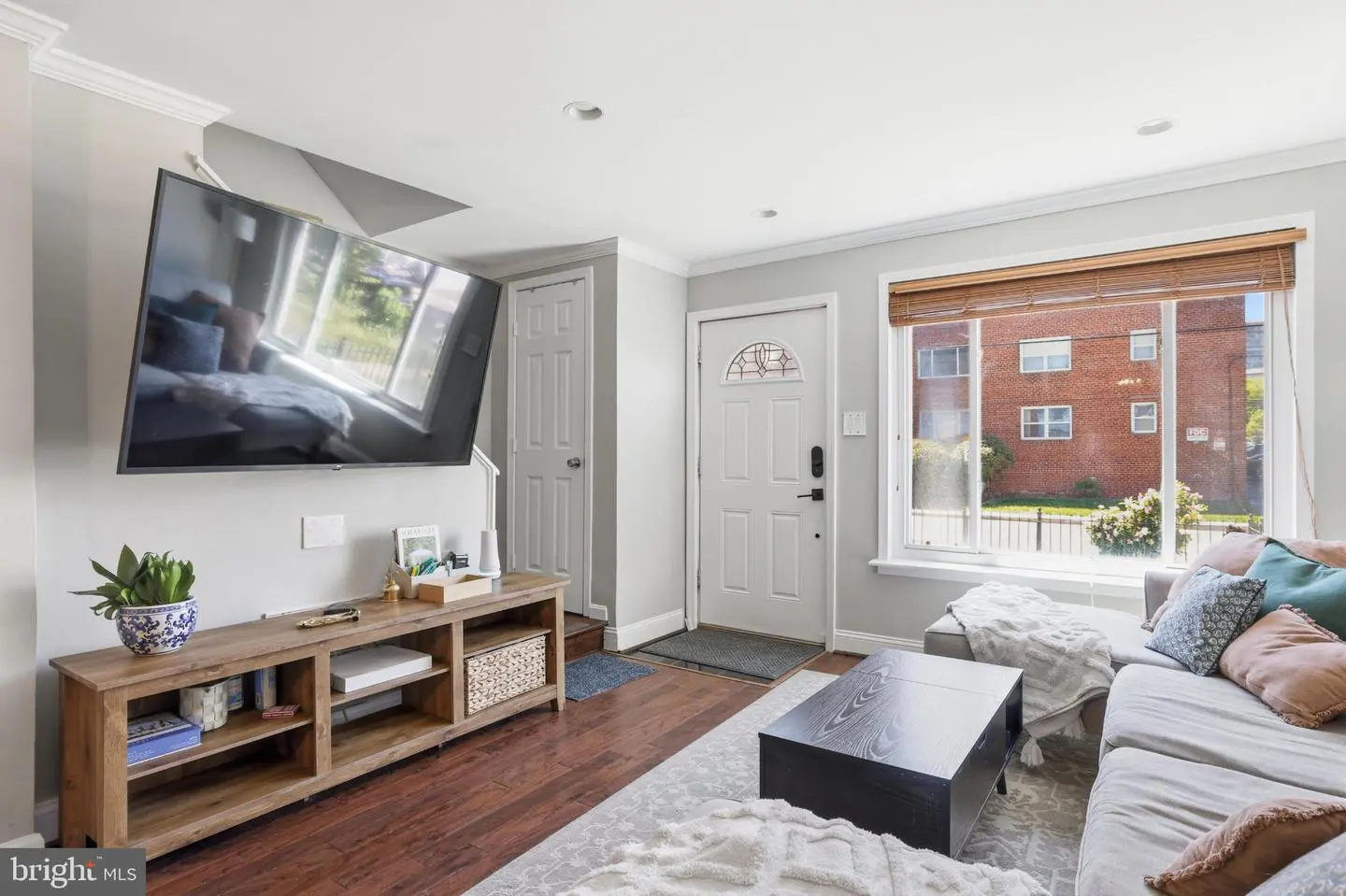 Living room with gray walls, wood floors, and a large window. A TV hangs above a wooden console table. A gray sofa faces a black coffee table.