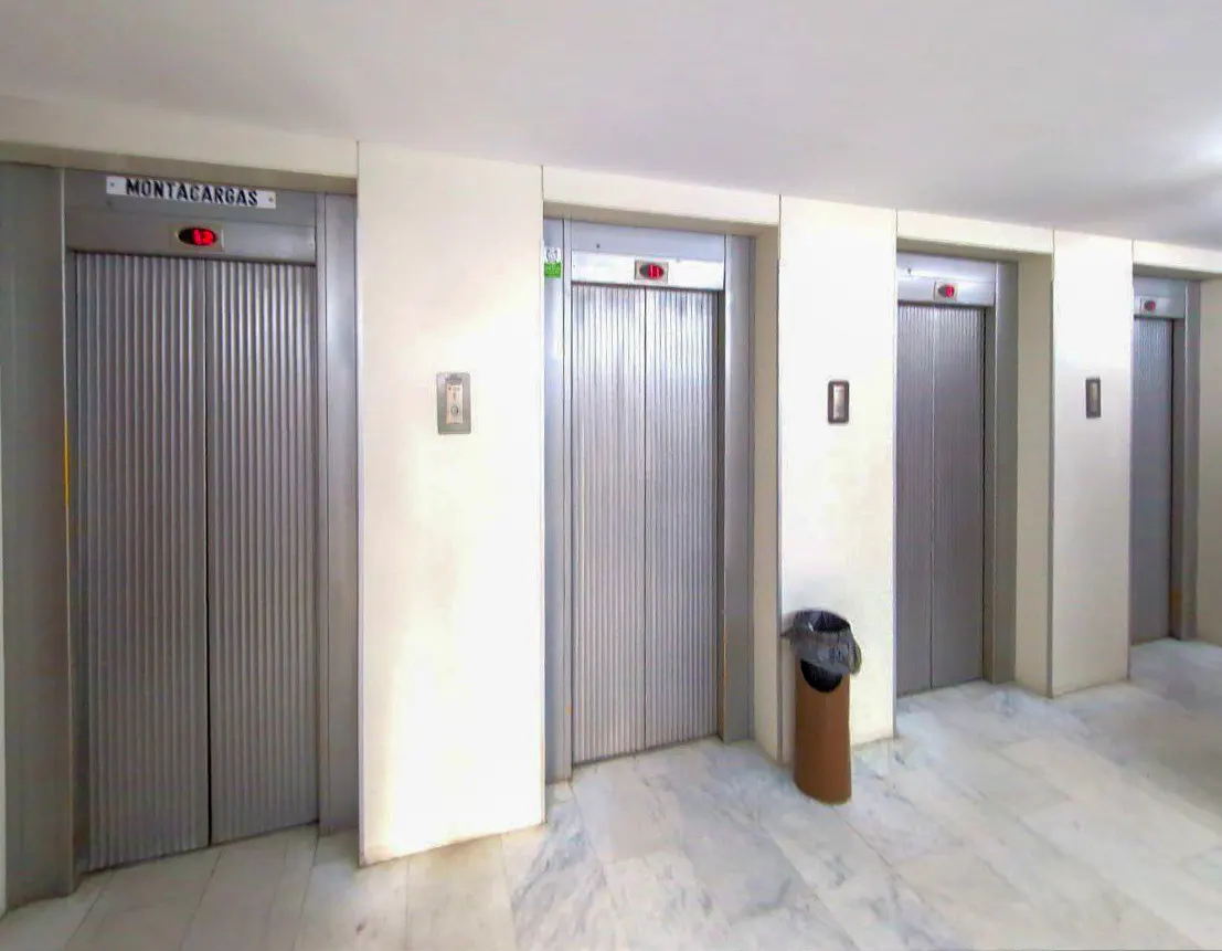 Four silver elevators in a white-walled lobby with a marble floor. A brown trash can sits near the second elevator.
