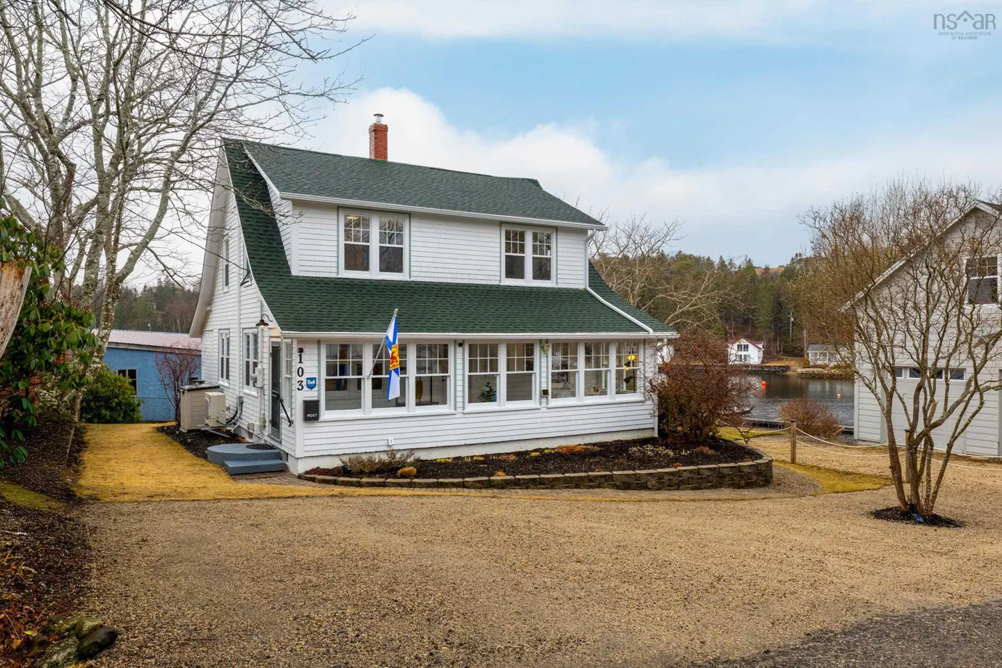 Two-story white house with a green roof and a Nova Scotia flag on the porch. A lake is visible in the background.
