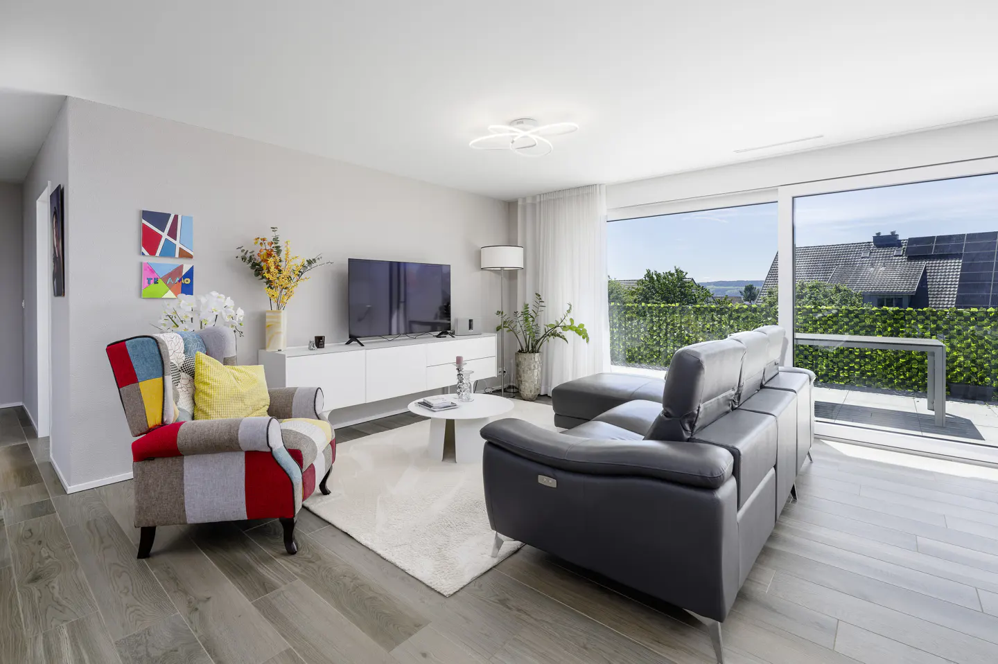 Modern living room with a grey sectional, colorful patchwork armchair, white media console, and large balcony windows.