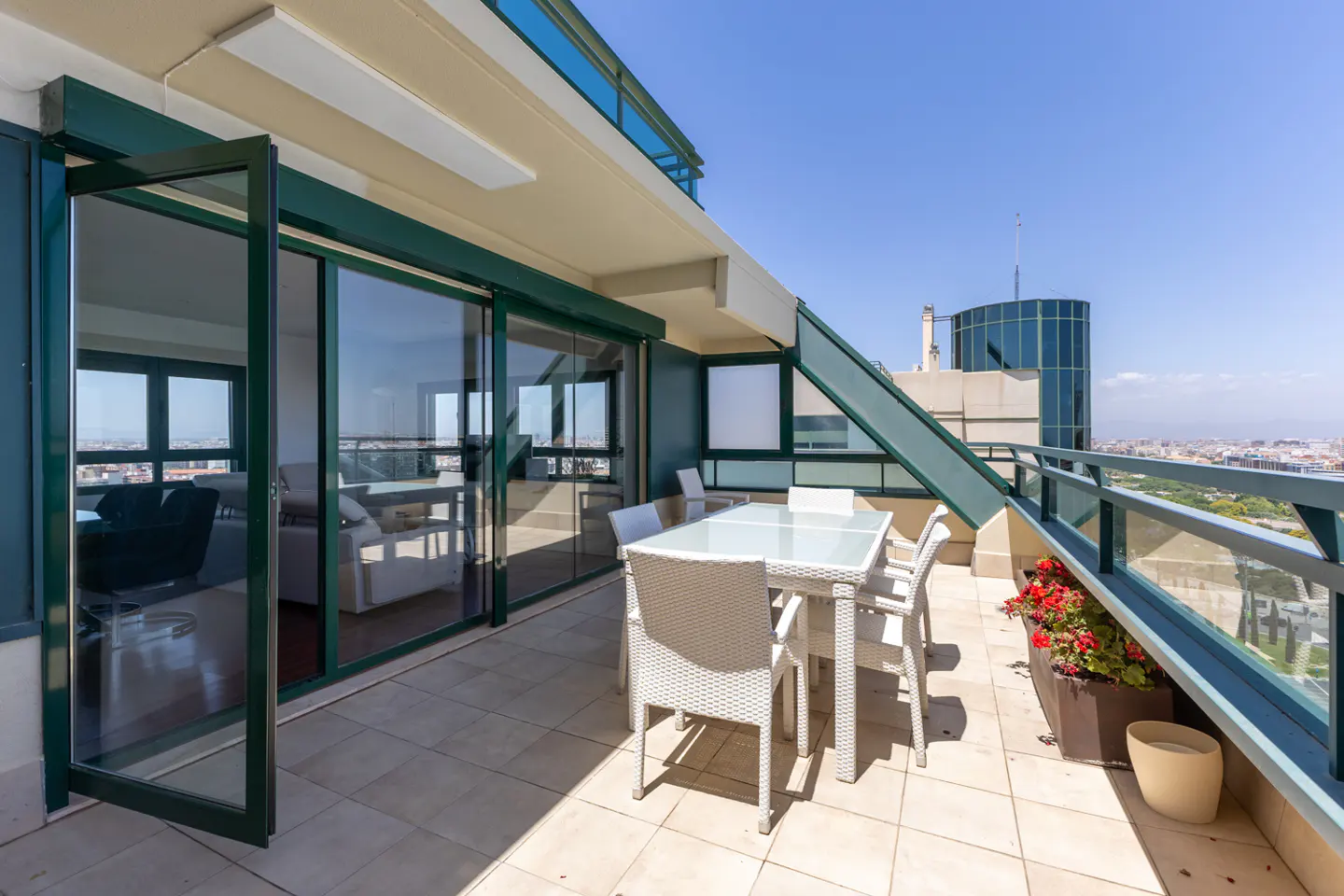 Modern balcony with white wicker dining set, glass railings, and city views under a clear blue sky.
