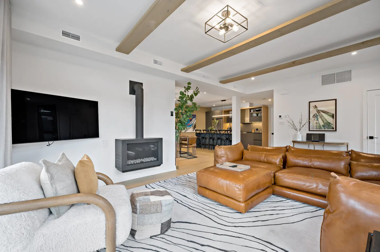 Living room with a brown leather sectional sofa, white armchair, and a modern fireplace. The ceiling has wooden beams and a square light fixture.