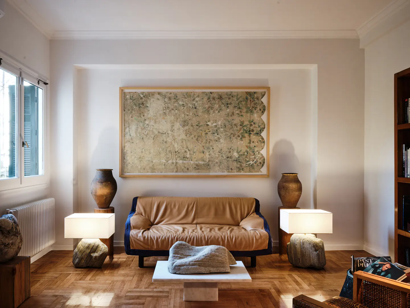 Living room with a tan leather couch, stone lamps, and a large abstract painting. Hardwood floors and white walls create a bright, modern space.
