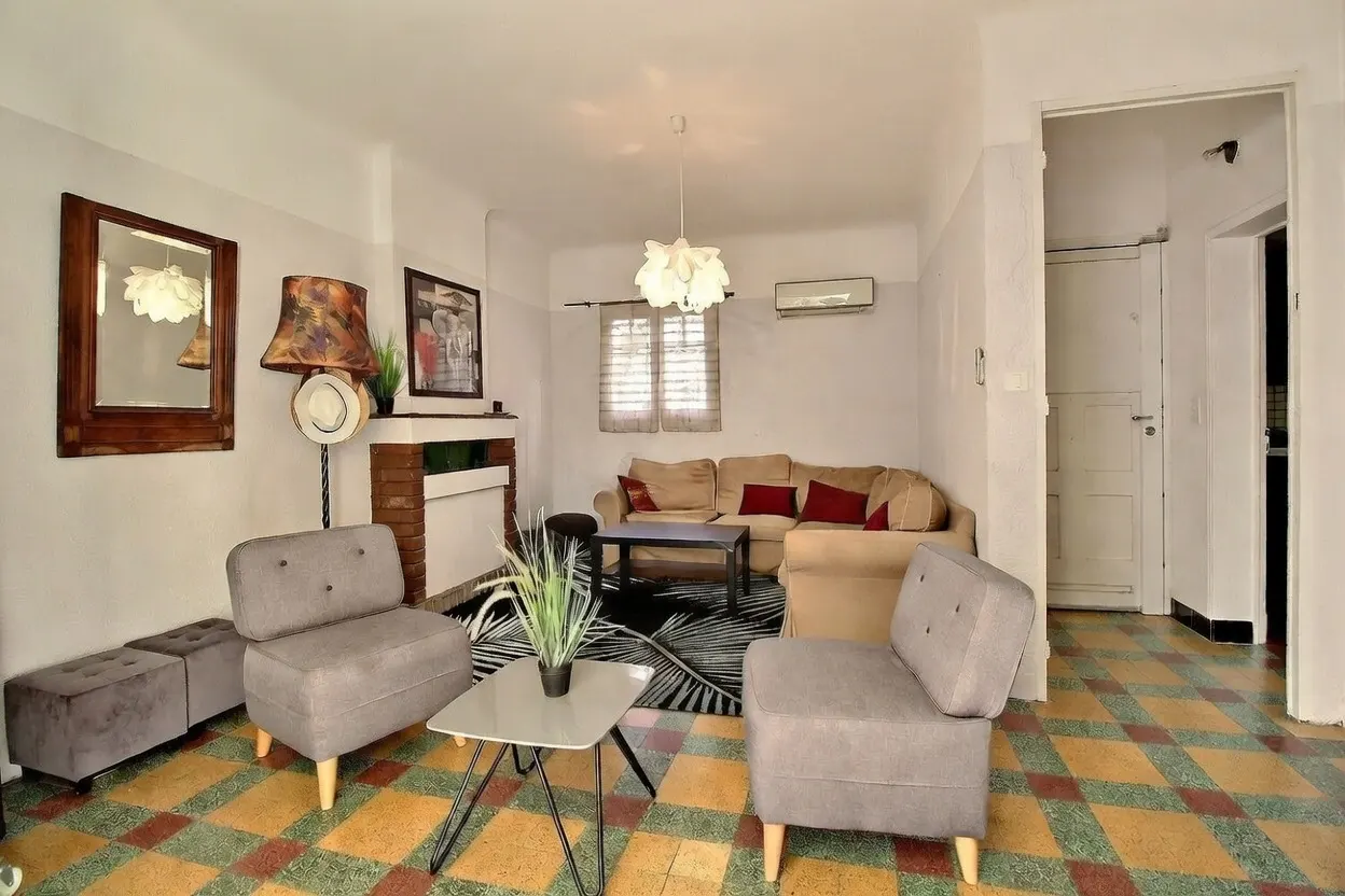 Living room with gray chairs, beige sofa, and patterned floor. A fireplace, mirror, and lamp add character to the bright, inviting space.