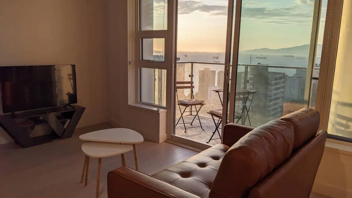 Living room with a brown leather couch, TV, and balcony with a city and ocean view at sunset.