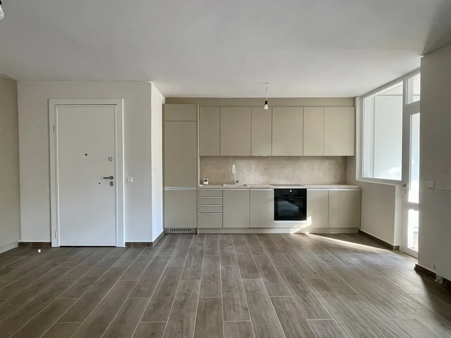 A bright, empty apartment with a kitchen, white door, and wood-look tile flooring.