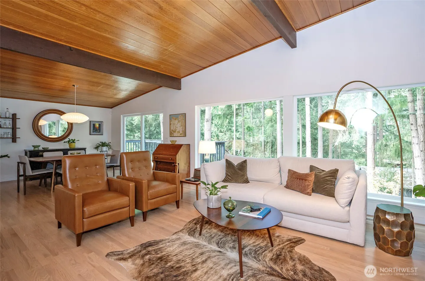 Bright living room with wood ceiling, white walls, and large windows overlooking trees. Leather chairs and a white sofa furnish the space.
