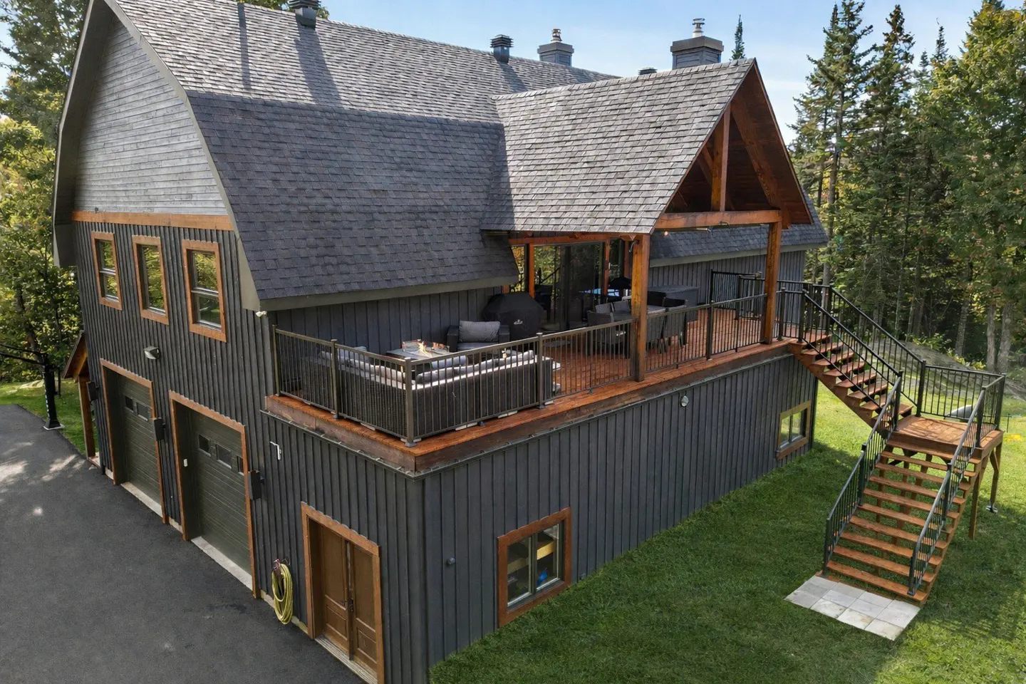 Exterior view of a modern gray barn-style house with a deck, outdoor furniture, and a staircase leading to the lawn.