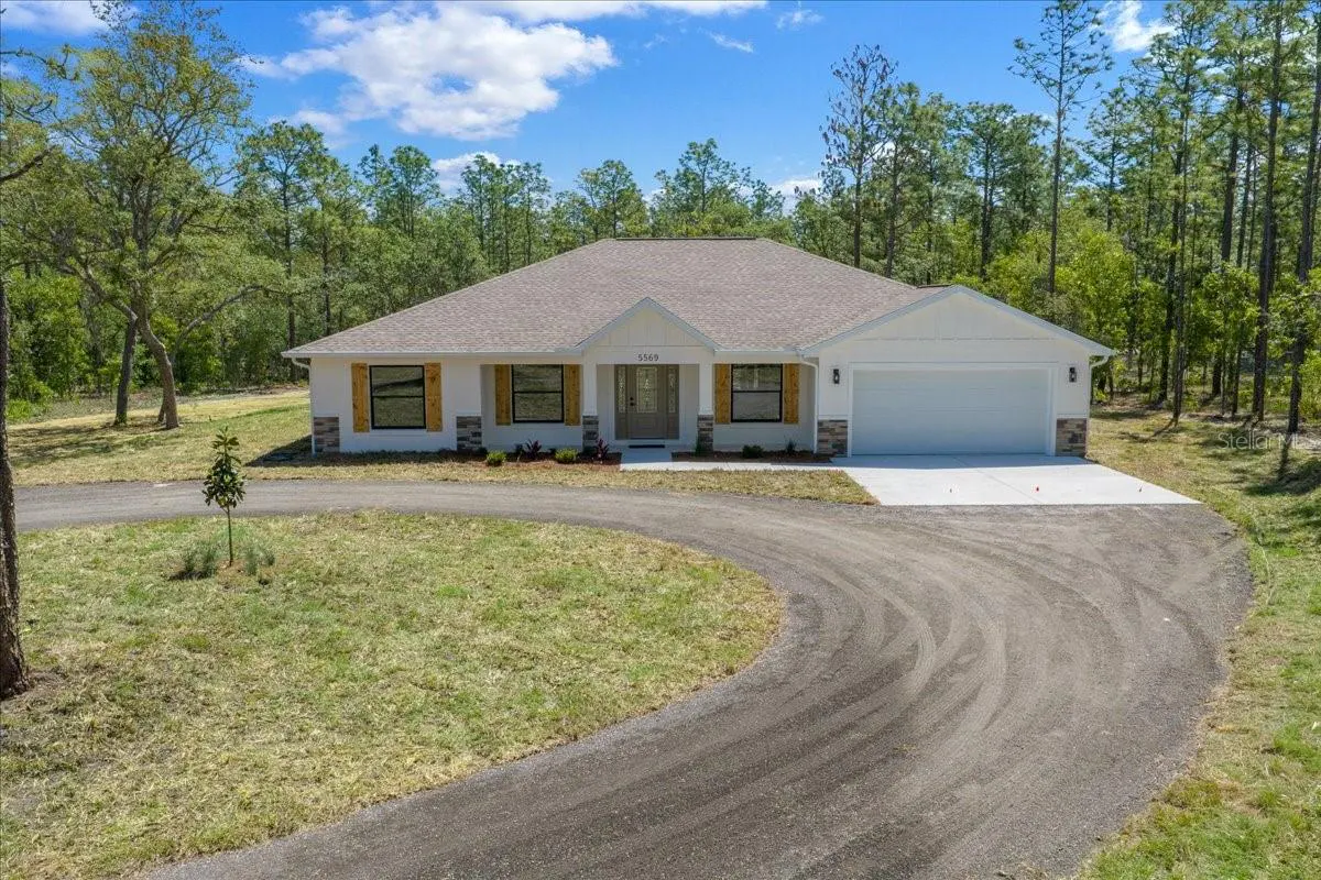A white single-story house with a gray roof and a curved driveway. The house has wood shutters and a two-car garage. Trees surround the property.