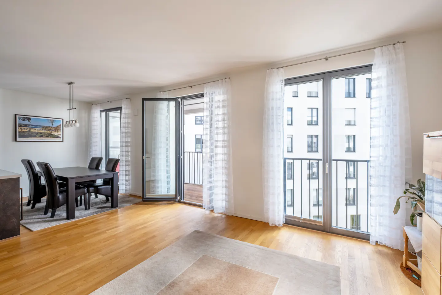 Bright, modern apartment interior with wood floors, a dining table, and balcony doors with sheer white curtains.